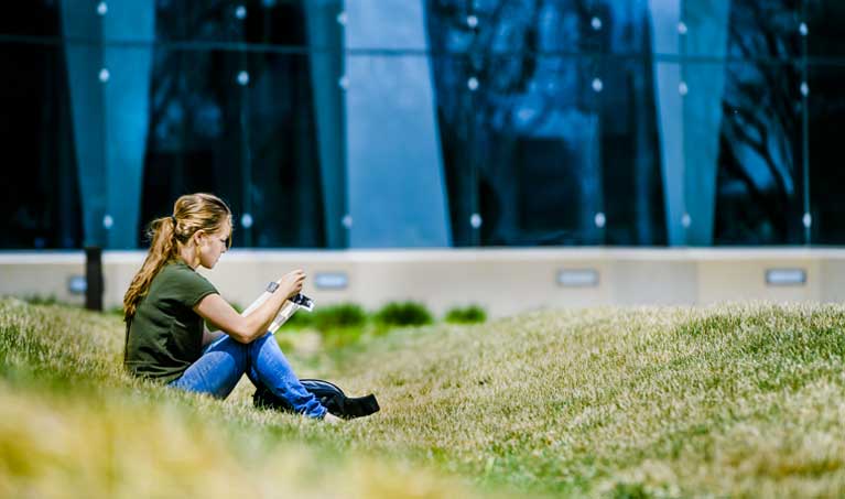 Woman sitting in the grass reading in front of the Gannon Building Glass