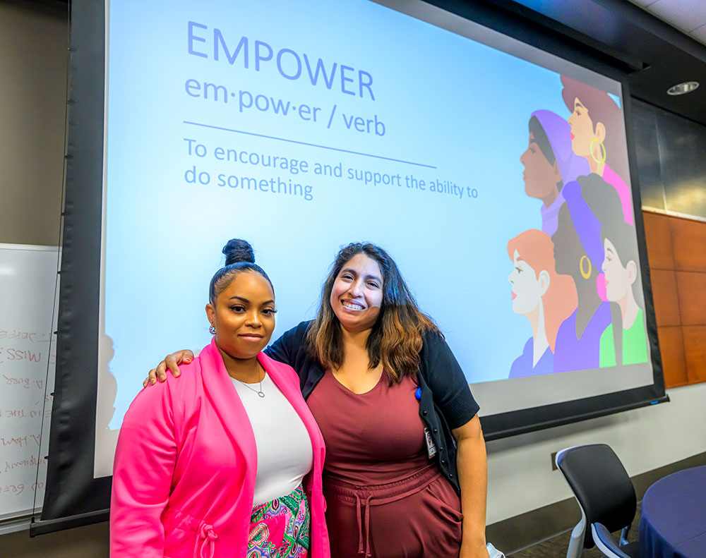 two women at the front of a class posing for a photo below a projector