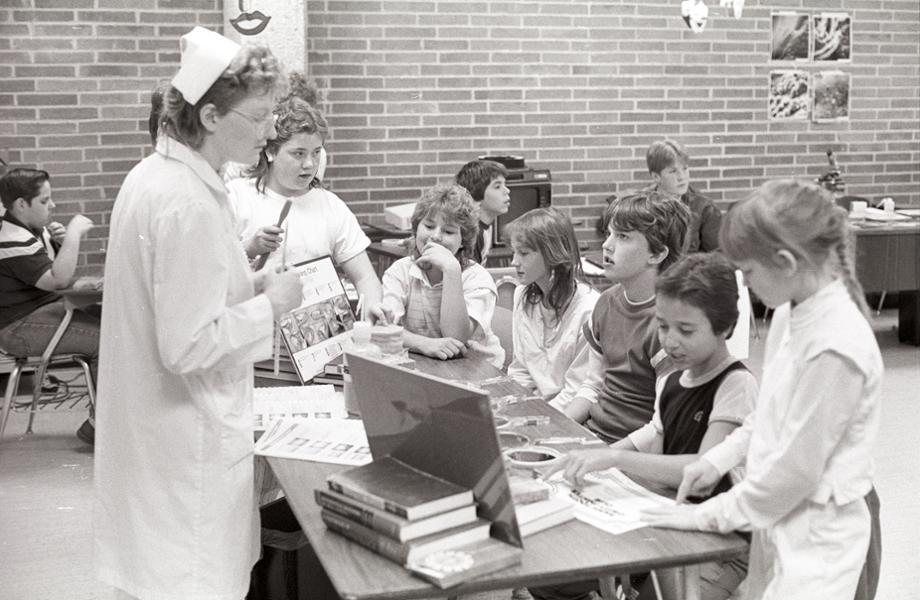 Young students stand at a table full of health related information and listen to the woman standing behind the table.