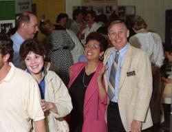 Two LCC staff members smile at the camera in the midst of a space full of students and other staff members.