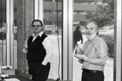 Two men eating ice cream cones in front of a wall of windows.