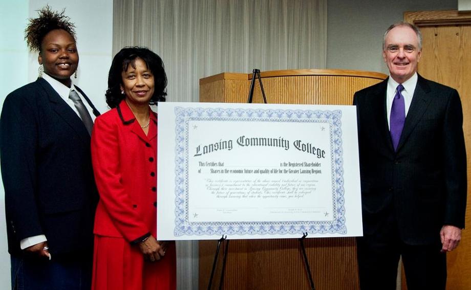 Three people stand around an oversized certificate on an easel.