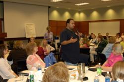 A Support Staff Day attendee stands at a table and speaks to the room using a microphone.